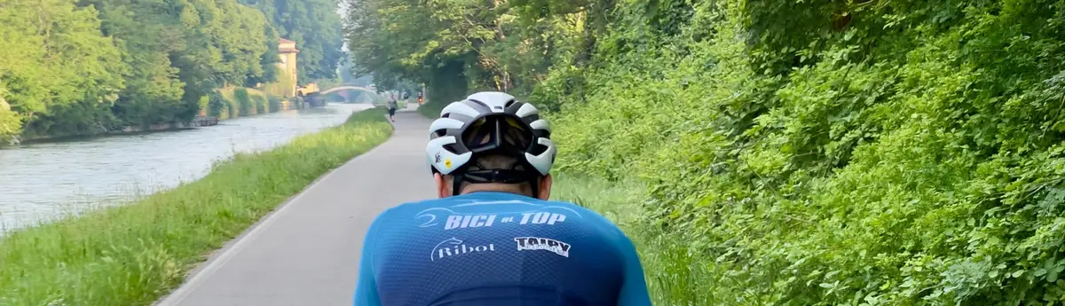 Un ciclista con la maglia col nome Ribot, di spalle, lungo l’alzaia di un canale nel verde