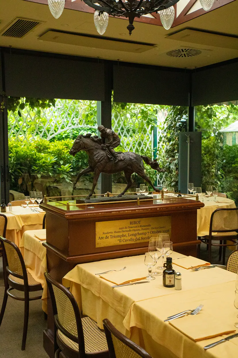 La veranda di Ribot a San Siro: al centro, fra i tavoli apparecchiati, il cavallo di bronzo col fantino sul suo piedistallo, e dietro le vetrate il giardino